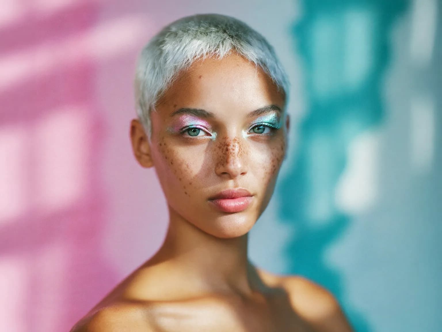 Portrait of a woman with silver-dyed buzz cut and freckles, wearing iridescent holographic eyeshadow, lit by soft window light with a pastel pink and teal gradient backdrop, ultra-detailed skin texture, contemporary beauty photography